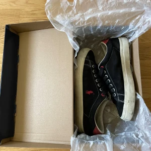 Svarta sneakers från Polo Ralph Lauren - Snygga svarta sneakers från Polo Ralph Lauren med röd logga på sidan och röda detaljer vid hälen. Skorna har vit sula, snörning och kontrastsömmar. Ovandelen är i mocka och de har en klassisk, rund tå. Perfekta för en stilren look. 
