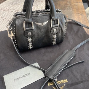 Zadig & Voltaire Sunny Nano Grai  - Liten svart handväska från Zadig & Voltaire i modellen Sunny Nano Grai med silverfärgade metalldetaljer, dubbla dragkedjor och nitar längs kanterna. Väskan har både handtag och axelrem, samt ikonisk vinge i metall framtill. Perfekt storlek för det viktigaste.