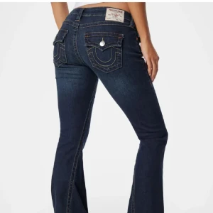 True Religion bootcut jeans mörkblå - Mörkblå bootcut jeans från True Religion med ikoniska kontrastsömmar och stora fickor bak med lock och silverknappar. Den är i storlek w28 och har låg midja och klassisk femficksdesign. Snygga detaljer och slitstarkt jeanstyg, perfekta för en cool och avslappnad look.