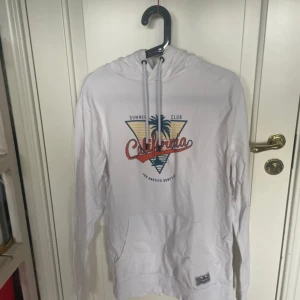 Vit hoodie från Denim Factory, M - Vit hoodie från Denim Factory med tryck framtill där det står 'California' och en palm. Tröjan har huva med snörning, känguruficka och långa ärmar. Perfekt för chill dagar och sommarvibbar. Materialet känns som bomull och passformen är normal.