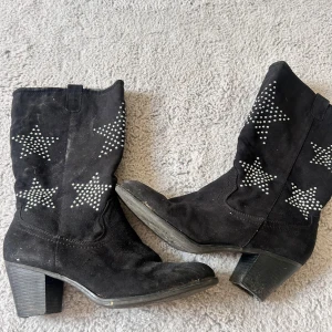 Svarta cowboy boots med stjärnor - Svarta cowboy boots i mocka med coola stjärnor av silvriga nitar på skaftet. Skorna har en bred klack och rund tå, perfekt för dig som vill sticka ut med din stil. Snygga detaljer och en klassisk western-vibe. Saknar en del på klacken men det syns bara underifrån