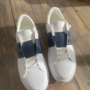 Valentino Garavani Open sneakers vit/blå - Snygga Valentino Garavani Open sneakers i vitt skinn med bred blå läderdetalj över vristen. Klassisk rund tå, vita snören och ikoniska nitar baktill på sulan. Perfekta för dig som vill ha en clean och lyxig look.