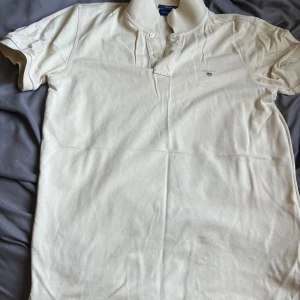 Beige GANT pikétröja med broderi - Snygg beige pikétröja från GANT i klassisk regular fit. Tröjan har korta ärmar, krage och knappar framtill. På bröstet finns ett broderat GANT-logo med sköld. Materialet är mjuk bomull som känns skönt mot huden. Perfekt för en chill och stilren look.