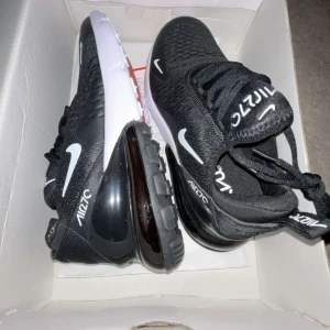 Nike Air Max 270 svart/vit strl 40 - Nike Air Max 270 sneakers i svart mesh med vita och orange detaljer. Snygg design med stor synlig Air-enhet i hälen, vit och svart sula samt orange undersida. Skorna har snörning och är perfekta för dig som gillar sportig och modern stil.
