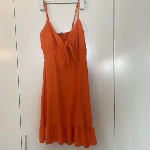 Orange sommarklänning med volang - Superfin orange kortklänning från New Collection med smala axelband, knyt framtill och volangkant nertill. Perfekt för varma dagar, lätt och luftig med smockad rygg för extra komfort. Snygg och enkel att styla till sneakers eller sandaler. Klänningen har ett litet ilägg i Bh delen. 