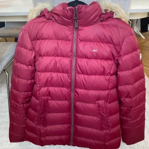Vinröd jacka Tommy Jeans S - Snygg vinröd pufferjacka från Tommy Jeans med fuskpäls på huvan. Jackan har dragkedja framtill, två fickor och quiltad design. Perfekt för vinter och kalla höstdagar 