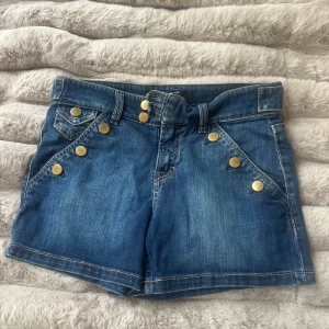 Blå jeansshorts med guldknappar Filippa K - Snygga blå jeansshorts från Filippa K med flera guldiga knappar fram och bak. Klassisk femficksdesign, normal passform och midwaist. Materialet är slitstarkt jeans och shortsen har coola detaljer som markerade sömmar och fickor med lock bak.