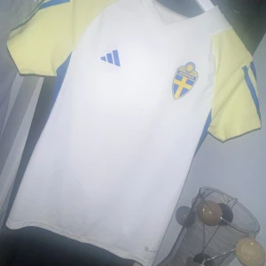 Svenska landslaget fotbollströja  - Säljer en vit fotbollströja från Adidas med Sveriges landslagsemblem på bröstet. Tröjan har korta, ljusgula ärmar och blå detaljer längs sidorna. Tillverkad i lätt och ventilerande funktionsmaterial, perfekt för träning. Aldrig använd.
