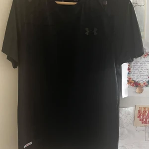 Svart Under Armour t shirt - Svart t-shirt från Under Armour med diskret logga på bröstet. Tillverkad i lätt och ventilerande syntetmaterial, perfekt för träning. Klassisk passform med korta ärmar och rund hals. Snygg och sportig stil som funkar till alla aktiva dagar. Regnbågig reflective. Passar storlek S då det är heat gear
