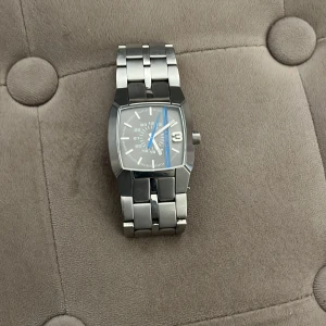 Diesel silverfärgad klocka med blå detalj - Stilren klocka från Diesel med fyrkantig urtavla, silverfärgat armband i metall och en snygg blå linje på urtavlan. Klockan har tydliga siffror och robust länkarmband som ger en cool och modern känsla.