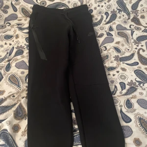 Svarta mjukisbyxor från Nike - Svarta mjukisbyxor från Nike med dragsko i midjan och diskret Nike-logga på låret. Byxorna har raka ben och en enkel, clean look. Perfekta för chill eller träning. Snygga detaljer med snörning och fickor på sidorna.