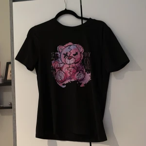 Svart t-shirt med rosa björntryck - Cool svart t-shirt med ett stort tryck av en rosa tecknad björn med blixt över ansiktet. T-shirten har rund hals och korta ärmar, perfekt för en avslappnad och trendig look. Passar dig som gillar unika och lekfulla motiv.