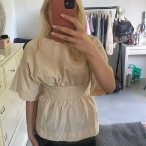 Beige blus med knytband i midjan - Säljer en beige blus med breda, korta ärmar och markerad midja med knytband i ryggen. Toppen har en luftig passform och lätt rynkade detaljer både upptill och nedtill. Perfekt för dig som gillar en stilren och trendig look.