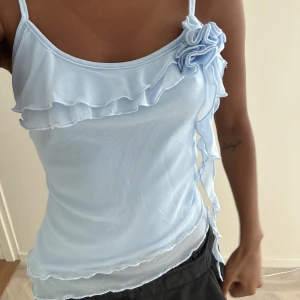 Ljusblått linne / top - Jättesött ljusblå snedskuret / asymmetriskt linne med blomma och volang / volanger.  Justerbara axelband.  Storlek XS   Nyskick! 💙🩵Använd köpknappen om du vill köpa💕 spårbar frakt via instabox eller PostNord 🌸