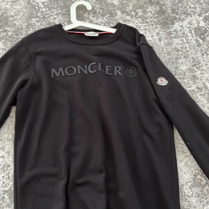 Moncler Hoodie - Säljer en Svart Moncler Hoodie denna varan är även i magnifikt skick. Har även ett Moncler märke på kanten vid armen och ett vid bröstet bredvid texten.