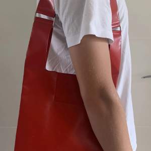 Röd axelväska i plast från Equipaje BCN. Knappt använd, men har fått ett litet märke på axelremmen, se bild. 35x24 cm är själva väskan, förutom remmen.