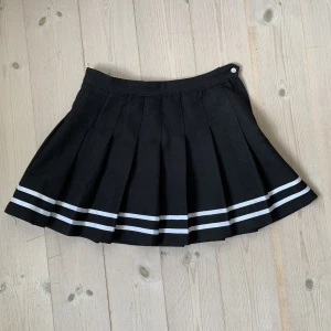 Svart tennis kjol - Svart veckad kjol med två vita påsydda ränder. Dragkedja och knapp på sidan. Styla som schoolgirl eller egirl!