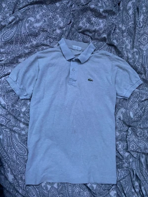 Lacoste piké  - Fin Lacoste piké i grå ganska bra skick. Nypris 1200kr. Väldigt litet hål på framsida men de syns inte om man inte kollar väldigt nära (Syns inte ens på bilden).