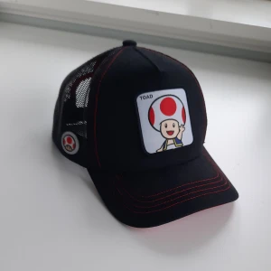 Super Mario Toad Truckercap - Jag säljer en fin Super Mario Bros Toad 2 Trucker Cap. Den är aldrig använd och är i juniorstorlek. 