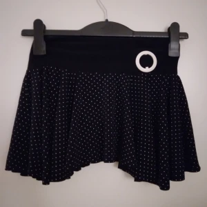 Svart prickig assymetrisk kort kjol med fastsydd trosa. Strlk: XS. Nyskick. - Svart prickig assymetrisk kort kjol med fastsydd trosa, bred resår i midjan samt vitt dekorativt spänne i ena sidan, från franskt fabrikat. Strlk: XS. Material: Syntet, troligen Polyester. Nyskick. Felfri.  