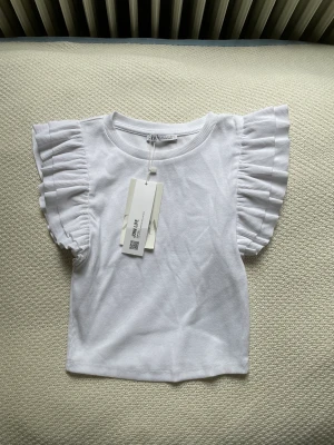 Volang T-shirt Zara - Säljer denna helt oanvända volang T-shirten ifrån zara. Aldrig använd med lappen kvar. Storlek S men passar även XS. Nypris: 119kr och jag säljer för 80kr
