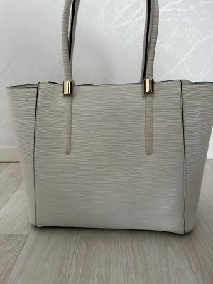 Väska  - En fin vit väska som har lite guldiga detaljer som även är märket tom leva den har vart använd ett par gånger ungefär 6-7 gånger den är väldigt fin och passar lite till vad som helst 