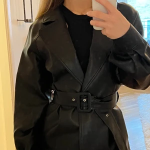 Skinnjacka med bälte - Svart skinnjacka(fakeläder) med bälte från NA-KD. Aldrig använd! Storlek EU 34. 300:- ex frakt. 