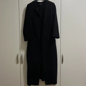 Tunn kappa - Tunn svart kappa med kortare ärmar, perfekt som lager-på-lager-jacka nu under hösten. Något figursydd modell med fickor på båda sidor. En liten diskret knäppning på insdan.