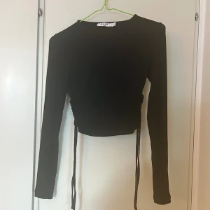 Svart ribbstickad långärmad tight topp - En svart ribbstickad, långärmad och tight topp som är liten i storleken. Är tunn i materialet och finns band som hänger ut längs sidorna som detalj. Funkar till vardags såväl som vid andra tillfällen. Använd endast en gång så är i mycket bra skick.