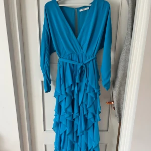 Blå maxiklänning  - Maxiklänning med volanger, slits, v-ringning och lång ärm från NA-KD. Storlek 32 men passar mig som normalt sett bär storlek XS. Använd endast en gång. Klänningen har ett knytbälte i midjan, samt både knappstängning och en dragkedja på baksidan.