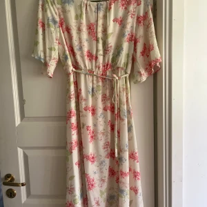 Blommig klänning - En vacker blommig klänning med korta ärmar och knytband i midjan. Klänningen har en ljus basfärg med blommönster i rött, grönt och blått. Den är tillverkad i ett lätt och luftigt material. har en öppning på framsidan
