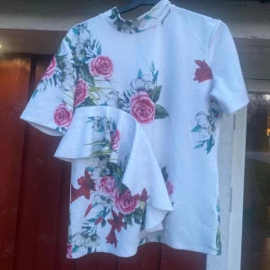 Fin blus med volang och blommönster. - Fin tröja/blus med vit bas och blommönster. Volang fram. Hög linning och knäppe bak. Mjuk och skön, 