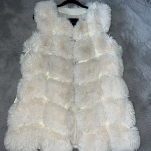 FAKE pälsväst - Krämvit figursydd mjuk pälsväst. 3 hakar på insida för att stänga igen. Två fickor på sidorna, varav en ficka som behövt limmas ihop (se sista bild)
