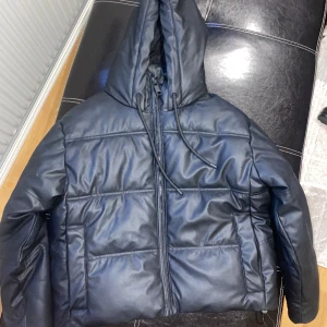 Dunjacka - En svart skinn aktig dunjacka. Köpt från Zara, org pris:900 kr. Den är kort, har luva och fickor för förvaring. Storlek S. Använd fåtal gånger, ser ut som ny.