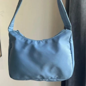 Ljusblå axelväska - En ljusblå axelväska med dragkedja. Väskan har en enkel och stilren design med en axelrem och är tillverkad i ett syntetmaterial. som det går att se på nästsista bilden har väskan en fläck som inte är så tydligt men kan vara bra att nämna