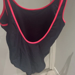 Svart linne från Champion - En svart baddräkt från Champion med färgglada Champion-logotyper tryckta på framsidan. Baddräkten har rosa kantband runt halsen och ärmhålen. Användt 1 gång och använder ej för har köpt ny baddräkt.