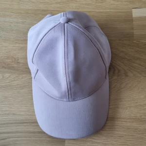 Ljusrosa keps från Lindex - En ljusrosa keps från Lindex med justerbar rem på baksidan. Kepsen har en klassisk design med en rundad skärm och en knapp på toppen.
