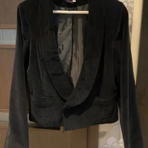 Svart kavaj - En kort svart kavaj med långärmad design. Kavajen har en klassisk krage och två fickor framtill. Materialet verkar vara av sammet, vilket ger en lyxig känsla.