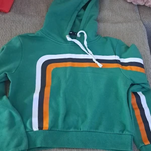 Grön hoodie med ränder - En grön hoodie med en vit dragsko i huvan. Tröjan har ränder i vitt, svart och orange på framsidan och på ena ärmen. Den har långa ärmar och en ribbad nederkant. använd 2 gånger bara. köpte för 300 säljer för 100