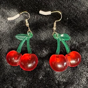 Nya örhängen m körsbär Cherries Cherry Pinup Kawai Rockabilly 50-tal 1950 Rock - Rasande söta örhängen med röda körsbär och gröna blad.  5 cm långa, 2,5 cm breda.   Krokar  - för dig som har hål i öronen.  Metall, plast.  Nya och oanvända. I originalförpackning.