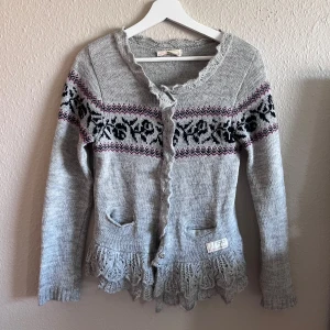 Grå stickad kofta med mönster - Säljer en superfin grå stickad kofta med ett svart och rosa mönster runt bröstet och axlarna. Koftan har långa ärmar och en söt volangdetalj längst ner. Den är i bra skick och perfekt för höst och vinter. Passar till både vardag och fest!