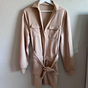 Beige klänning med knytband - Säljer en superfin beige klänning i storlek M. Den har långa ärmar och ett knytband i midjan som ger en snygg passform. Klänningen har också två fickor på bröstet och en krage som ger en stilren look. Perfekt för både vardag och fest!