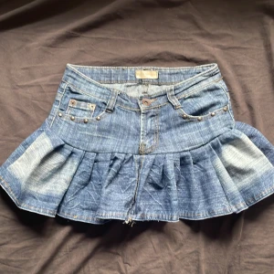 Jeanskjol med volang och broderi - Säljer en superfin jeanskjol med volangdetaljer och broderi på baksidan. Kjolen är kort och har en snygg, sliten look. Perfekt för sommarens alla fester! Den har också små nitar och en dragkedja fram. Passar perfekt till en casual outfit eller för att klä upp sig lite extra.