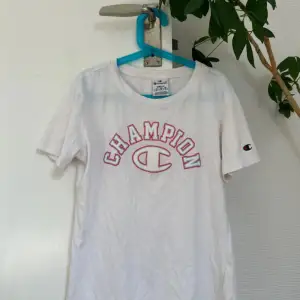 Säljer en vit t-shirt från Champion i storlek S. Den har korta ärmar och ett stort Champion-tryck på framsidan i rosa och vitt. Perfekt för en avslappnad och sportig look. T-shirten är i bra skick och redo för en ny ägare!