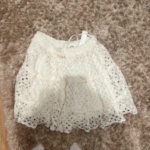 Vit spetskjol från Zara - Säljer en superfin vit spetskjol från Zara i storlek XS. Kjolen har ett vackert broderat mönster och en volangkant som ger den en söt och romantisk look. Den är perfekt för sommaren och kan bäras både till vardags och fest. Kjolen har en elastisk midja för en bekväm passform och en liten snörning med en söt snäcka som detalj. Den är i nyskick och knappt använd. Perfekt för att skapa en chic och somrig outfit! Nypris cirka 500🌸