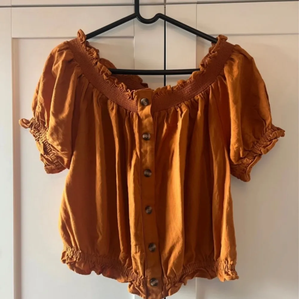 Säljer en superfin orange off-shoulder topp i storlek L. Den har en söt rynkad design med knappar framtill och elastiska ärmslut och nederkant. Perfekt för sommaren och kan stylas både uppklätt och nedklätt. Materialet känns lätt och luftigt, vilket gör den idealisk för varma dagar. Köp den nu och ge din garderob en färgklick! 🌞. Toppar.