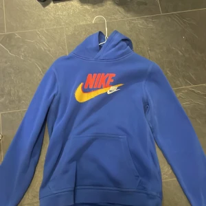 Blå hoodie från Nike - Säljer en snygg blå hoodie från Nike i storlek XL. Den har en stor Nike-logga i rött och gult på framsidan och en mindre logga på baksidan. Hoodien är perfekt för höst och vinter, och den är superbekväm med en normal passform. Materialet känns som bomull och är riktigt mjukt. Perfekt för både träning och vardag!
