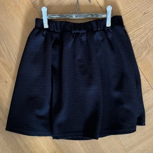 Svart kjol - En svart kjol med elastisk midja och texturerat tyg. Kjolen har en enkel och stilren design. Kjolen har även bälteshällor vilket gör att du enkelt kan bära ett skärp.