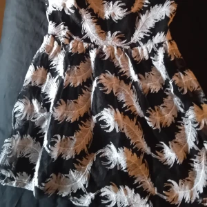 Svart klänning med vita fjädermönster - En elegant svart klänning med vita fjädermönster. Klänningen har en kort längd och är ärmlös, vilket gör den perfekt för festliga tillfällen.