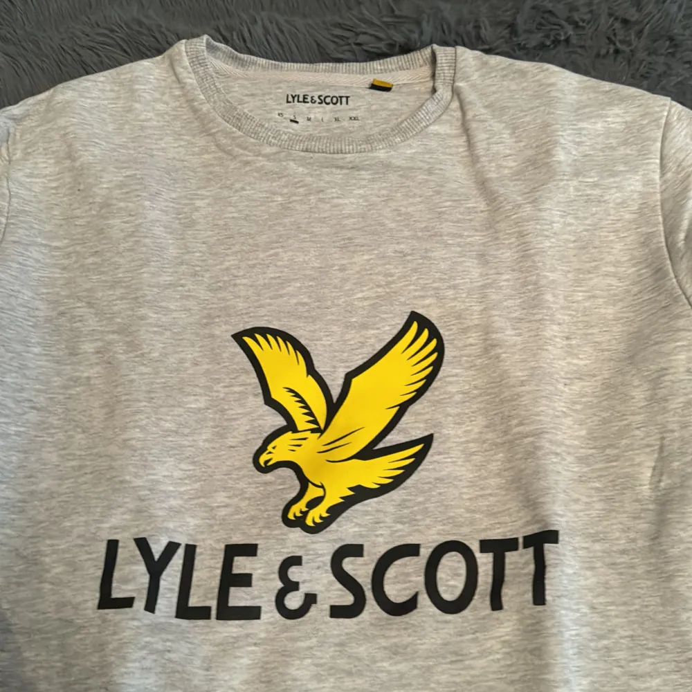 En bekväm grå sweatshirt från Lyle & Scott med ett stort gult fågeltryck och varumärkesnamnet på framsidan. Tröjan har långa ärmar och ribbstickade muddar vid ärmslut och nederkant.. Tröjor & Koftor.